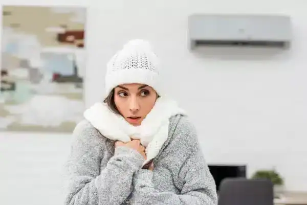 A woman dressed in warm winter clothing, including a knit hat, scarf, and sweater, stands indoors shivering from the cold. An air conditioner installed by an HVAC Contractor North Attleboro is mounted on the wall behind her.