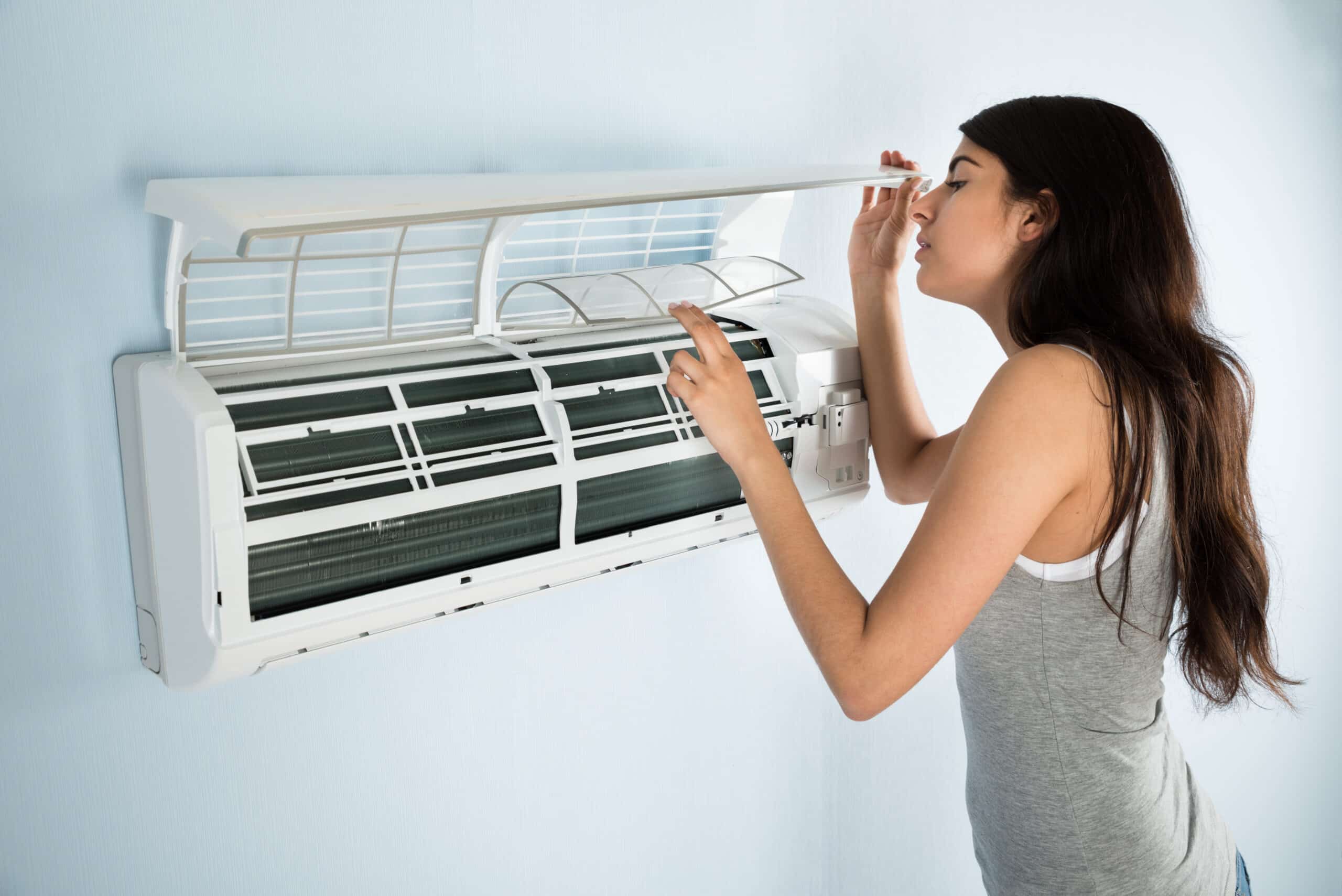A woman in a gray tank top inspects the inside of a wall-mounted air conditioner with the front cover lifted, against a light blue wall—an example of routine AC maintenance before calling an HVAC Contractor North Attleboro, MA.