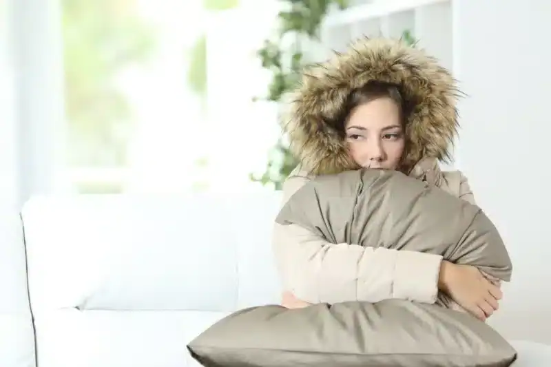 A woman wearing a thick winter coat with a fur-lined hood sits indoors on a white couch, hugging a gray pillow and appearing cold—clearly in need of AC Services North Attleboro to restore comfort.