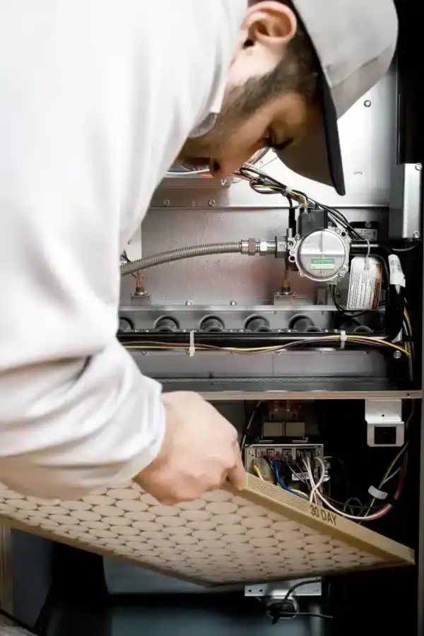 A person wearing a cap is installing or replacing an air filter in a furnace, showcasing the expertise of an HVAC Contractor North Attleboro, with visible wires, gas line, and mechanical components inside the unit.