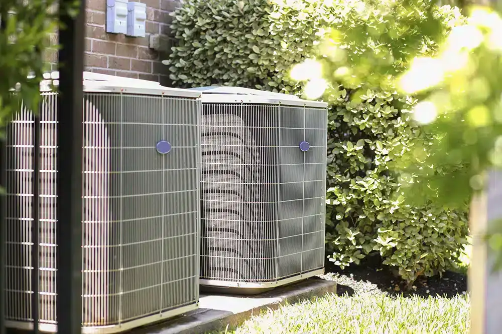 Two outdoor air conditioning units sit on a concrete pad beside a brick wall, surrounded by greenery in a sunlit yard—an example of quality AC installation North Attleboro residents can rely on.