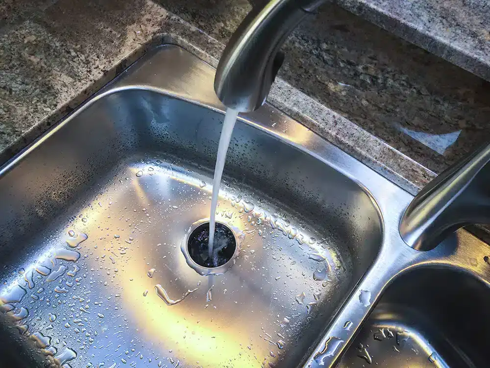 A kitchen faucet runs water into a stainless steel sink, with droplets scattered around the sink basin and counter—perfect for homes serviced by trusted HVAC Contractor North Attleboro, MA, ensuring comfort year-round.