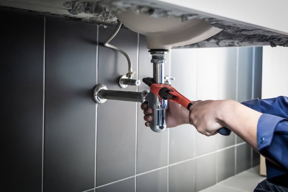 A person uses a red pipe wrench to tighten plumbing under a bathroom sink, surrounded by black tiles—ideal for those seeking expert HVAC Contractor North Attleboro services for all their home maintenance needs.