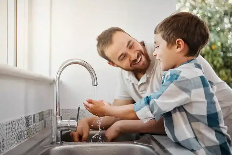 Father and son washing hands together at a kitchen sink. The boy, wearing a blue and white checkered shirt, holds his hands under running water while the father smiles, assisting him.