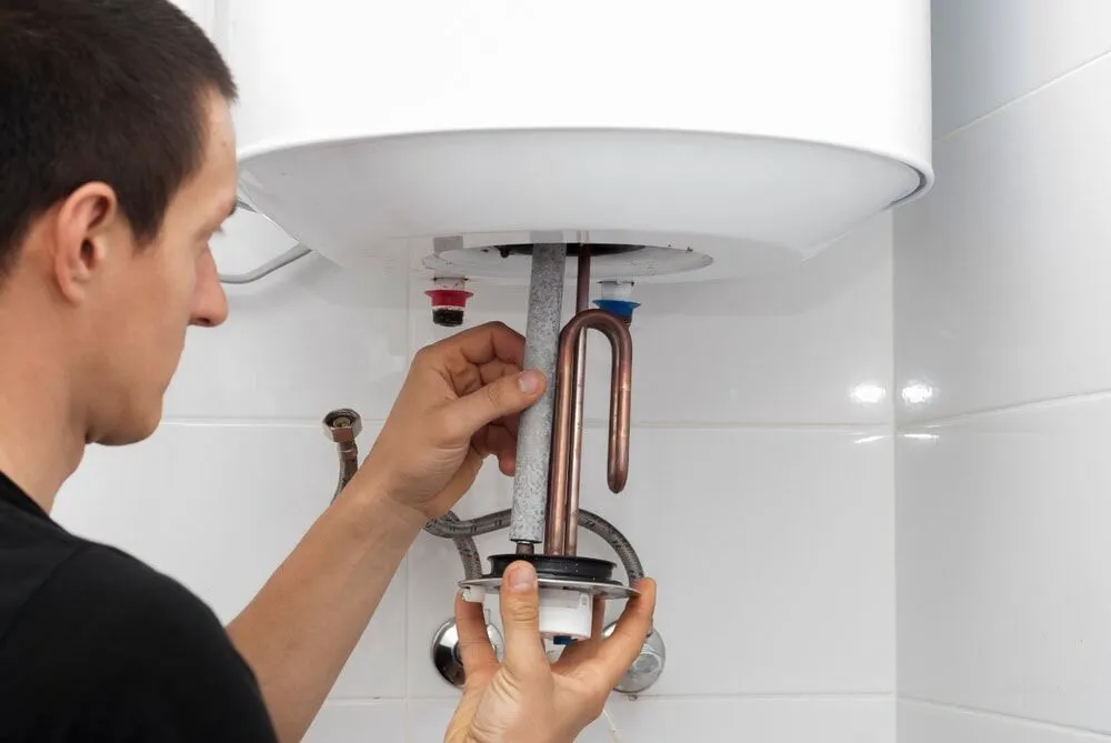 A person is repairing or installing a water heater in MA, holding a heating element and parts under the tank, with visible plumbing and pipes on a white tiled wall.
