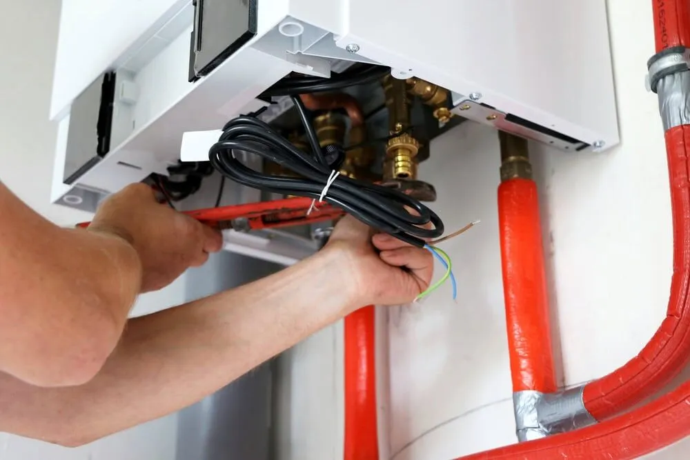 A person’s hands use tools to repair or install a wall-mounted boiler with exposed pipes and wires, including red insulated pipes connected beneath the unit—typical work for an experienced HVAC Contractor North Attleboro.