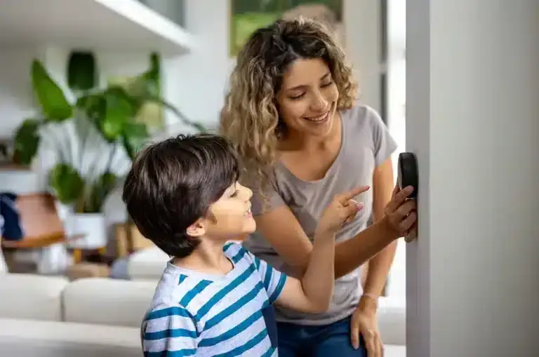 A woman and a smiling boy stand indoors, both looking at and touching a smart thermostat mounted on the wall—perfect comfort made simple with AC Services North Attleboro. Bright natural light and green plants fill the background.
