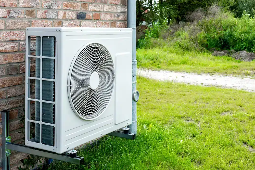 A white air conditioning unit is mounted on an exterior brick wall, with green grass and trees in the background. For expert AC installation North Attleboro trusts, choose a reliable HVAC contractor for quality service.