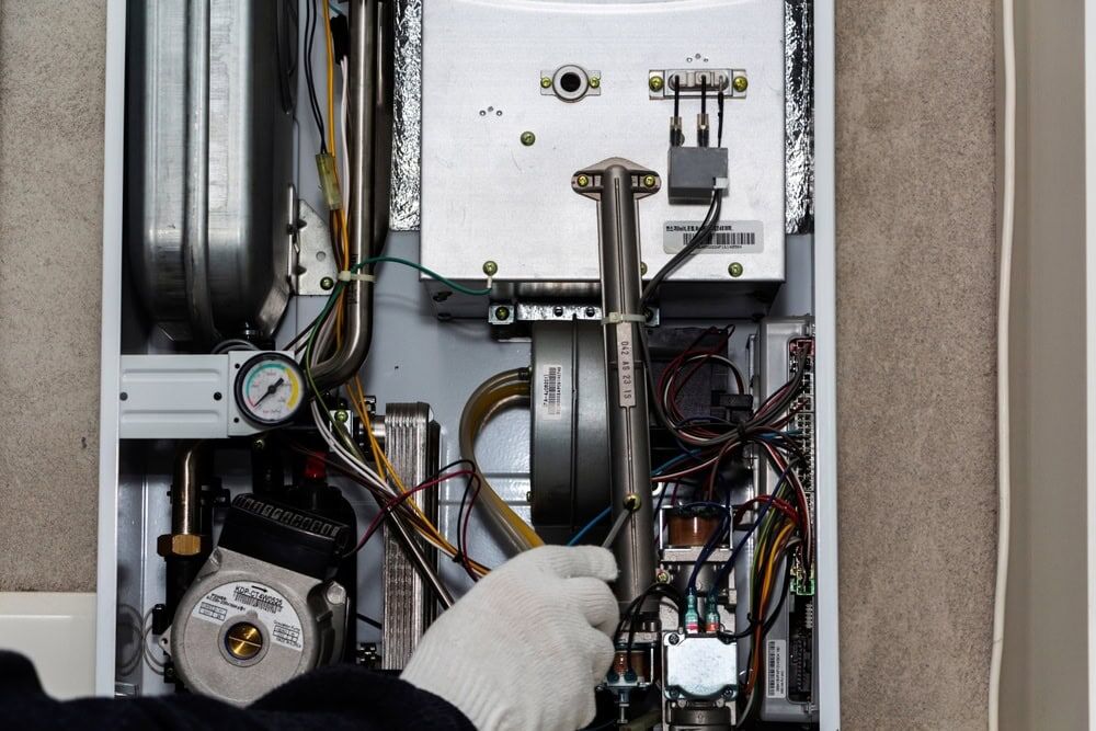 A gloved hand is adjusting wires inside an open gas boiler, highlighting various components, a pressure gauge, and intricate parts—showcasing the expertise of an HVAC Contractor North Attleboro.