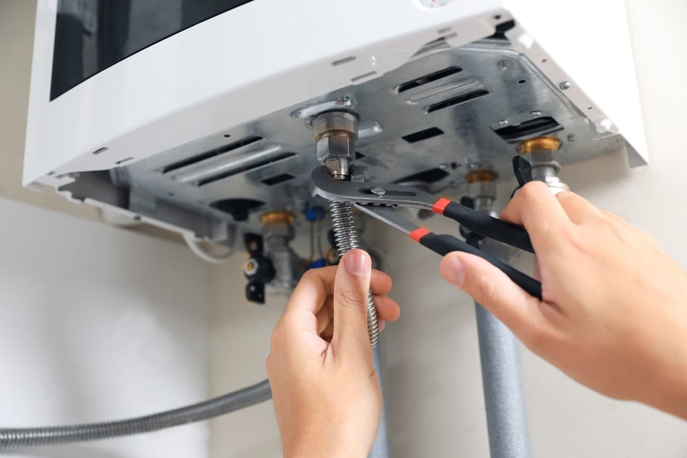 Hands using pliers to tighten a metal pipe connection on the underside of a wall-mounted water heater, with several visible pipes and fittings—professional service like AC Installation North Attleboro ensures secure connections and reliable operation.