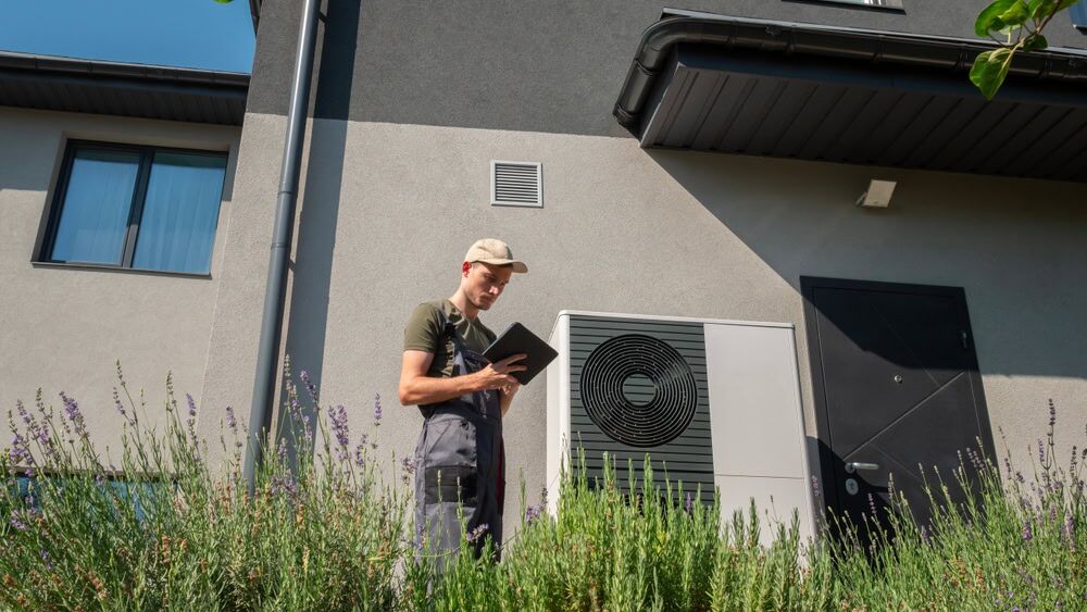 A technician in overalls and a cap stands outside a modern building, holding a tablet and inspecting an outdoor heat pump unit near flowering plants—showcasing expert AC Installation North Attleboro, MA services.