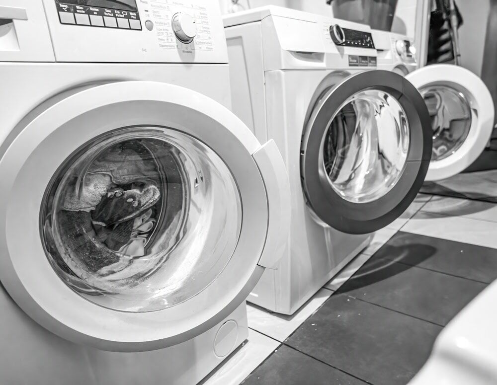 Two front-loading washing machines sit side by side in a North Attleboro laundry room. The black and white image reflects the surroundings, hinting at the need for reliable HVAC Contractor North Attleboro services to keep things running smoothly.