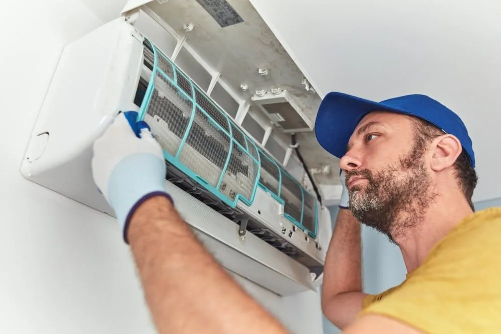 A man wearing a blue cap and gloves is cleaning or repairing a wall-mounted air conditioner, lifting the front cover and inspecting the filters—typical of an experienced HVAC Contractor North Attleboro, MA.