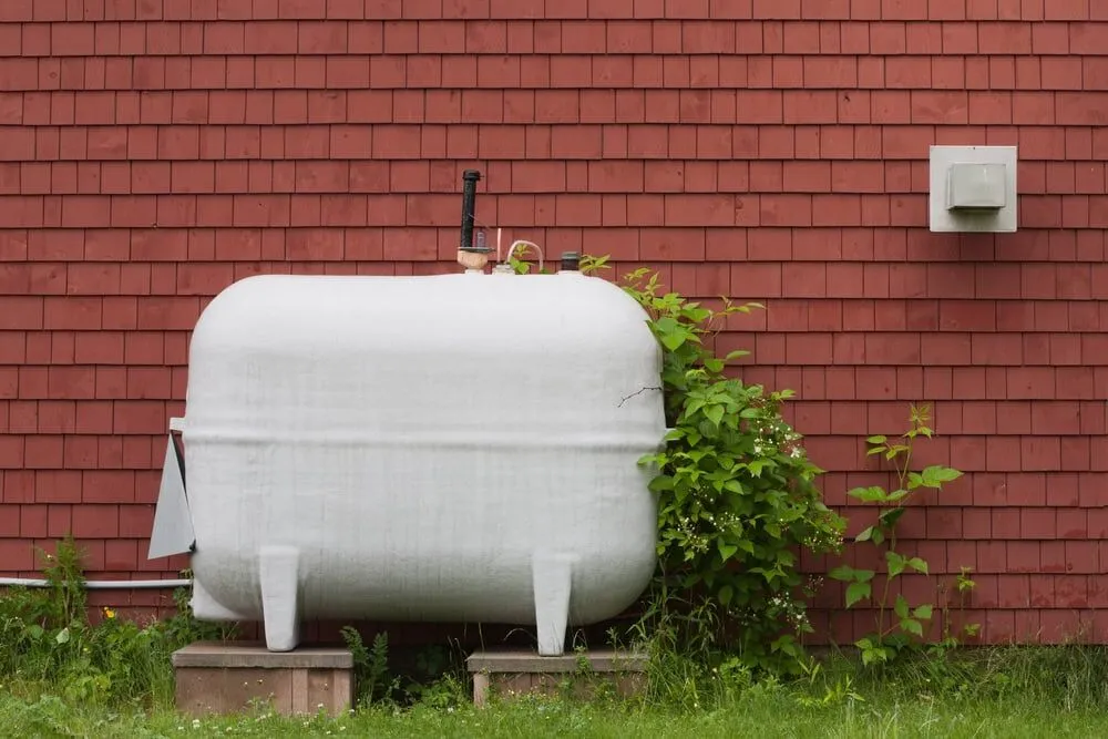 A large white propane tank stands on concrete blocks in front of a red shingled wall, surrounded by green grass—a typical sight for properties serviced by an HVAC Contractor North Attleboro, MA.