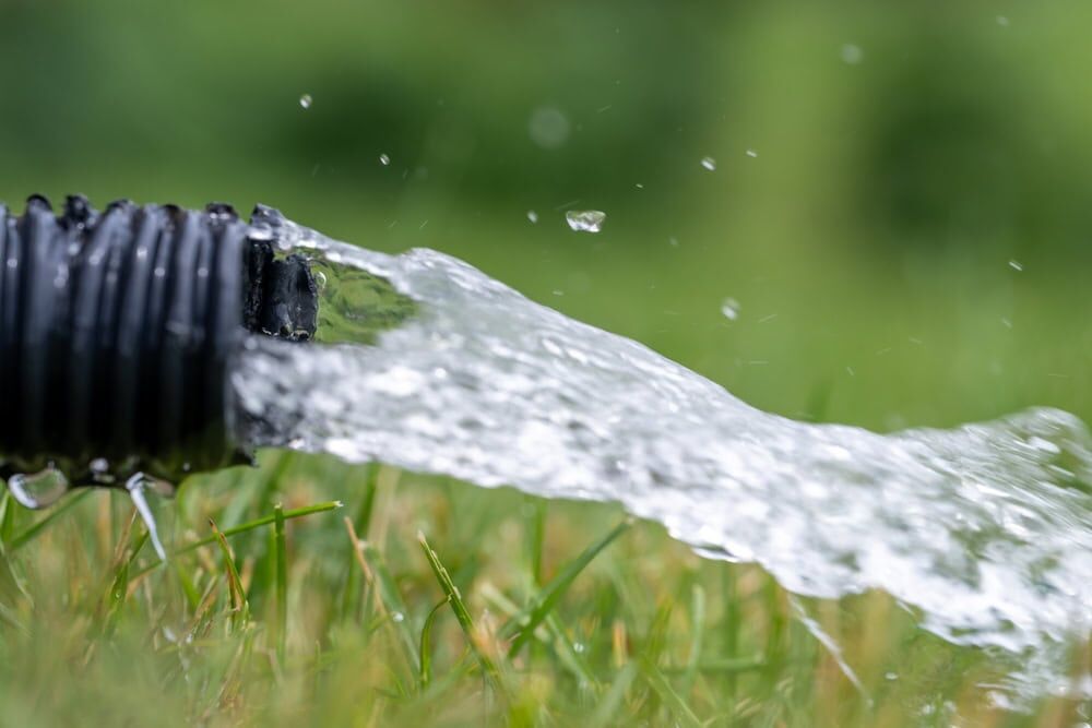 Close-up of water gushing out of a black garden hose onto green grass, with droplets and splashes in the air. The background is blurred—a refreshing scene for any AC Installation North Attleboro project on a warm day.