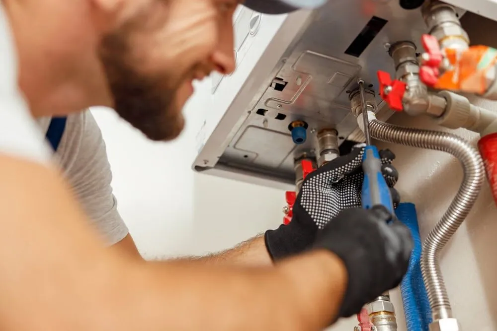 A plumber wearing gloves uses a wrench to tighten a pipe connection beneath a water heater, focusing on repair or maintenance work—showcasing the expertise you’d expect from an HVAC Contractor North Attleboro trusts. Various pipes and fittings are visible.