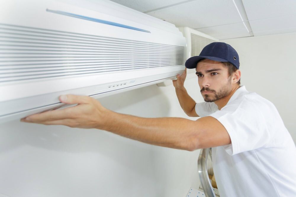 A man wearing a navy cap and white shirt is installing or repairing a wall-mounted air conditioner indoors, carefully adjusting the front panel—showcasing expert AC Services North Attleboro.