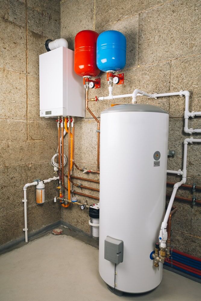 A boiler room in MA with a white water heater tank, two expansion tanks (one red, one blue) above, and various copper and plastic pipes linked to a wall-mounted control unit. The insulated walls ensure energy efficiency. Trusted by AC Services North Attleboro.