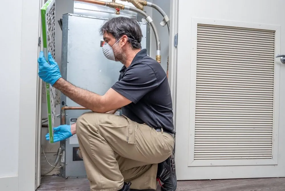A man wearing gloves and a face mask kneels while inspecting or replacing an air filter in an HVAC unit inside a utility closet—professional service from an experienced HVAC Contractor North Attleboro.