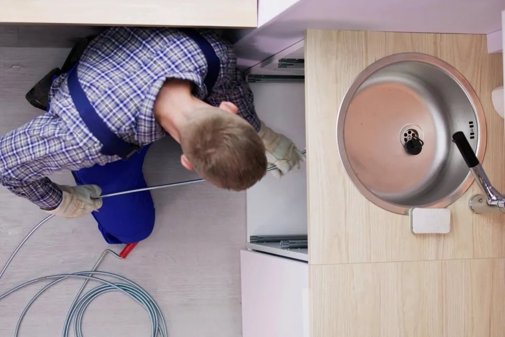 A plumber in gloves and a plaid shirt uses a drain snake to unclog pipes under a kitchen sink, viewed from above. Coiled cable and tools are spread out on the floor nearby, ready for any plumbing or AC services North Attleboro residents may need.