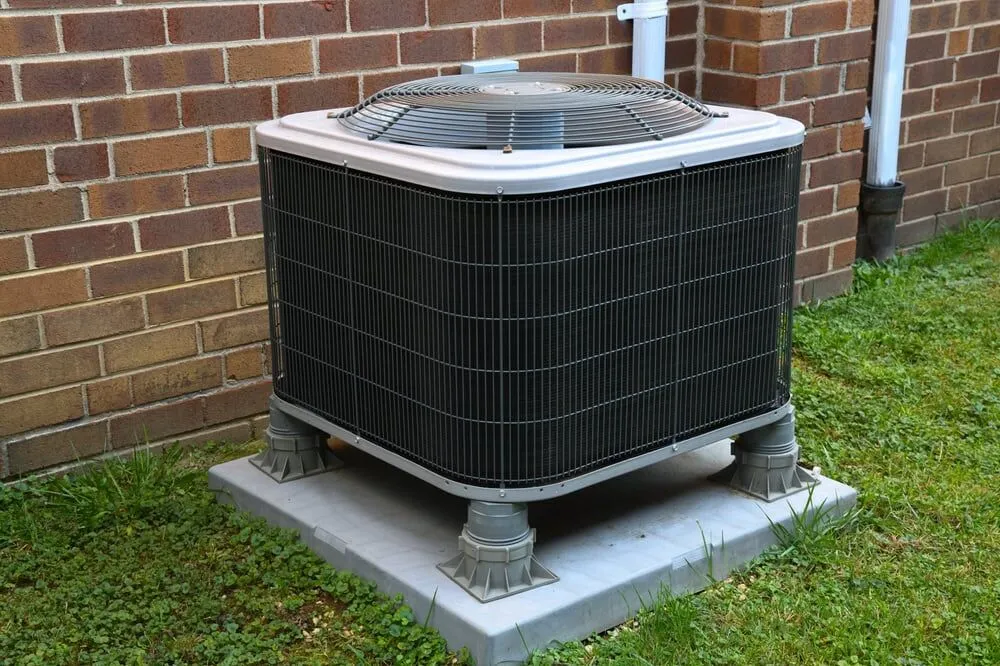 A central air conditioning unit sits on a concrete pad outside a brick building, surrounded by grass and positioned near utility pipes—perfectly maintained by a trusted HVAC Contractor North Attleboro.