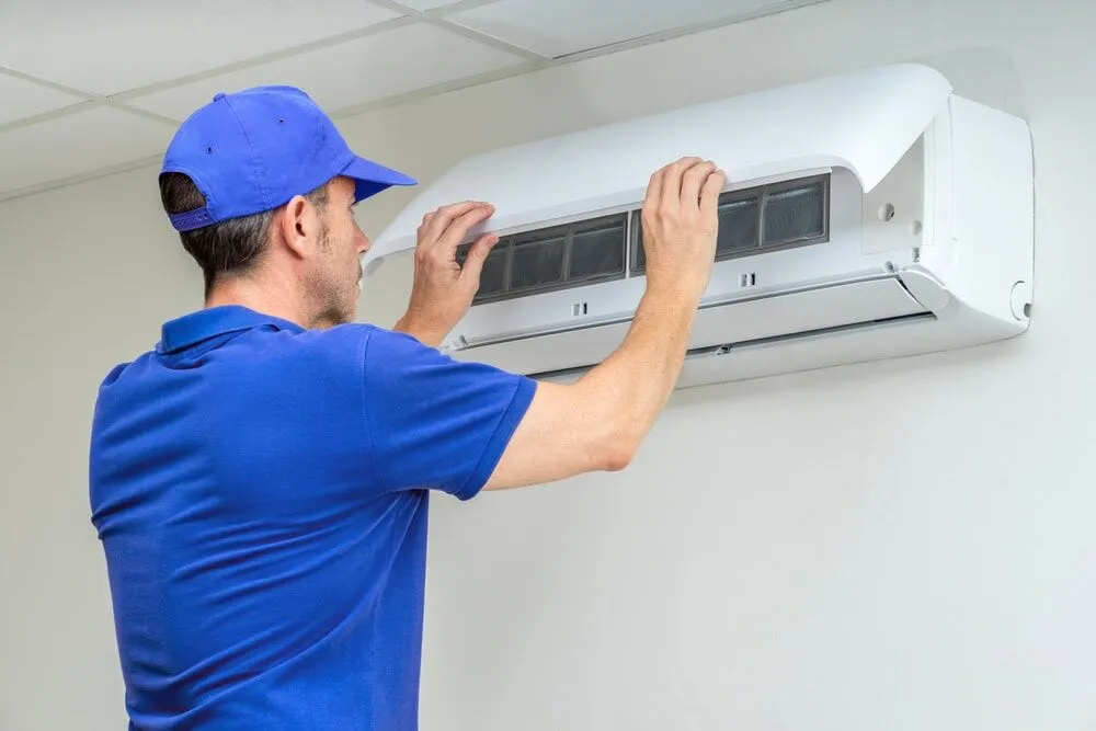 A technician in a blue uniform and cap is inspecting or repairing a wall-mounted air conditioner unit, with the front panel open—showcasing expert AC Services North Attleboro, MA.