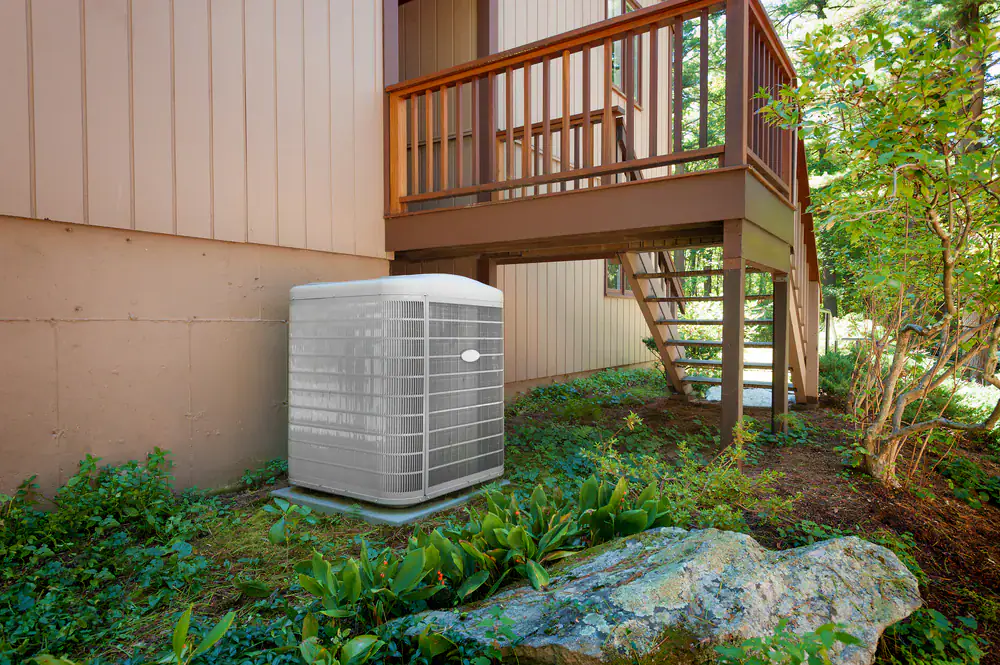 A central air conditioning unit, professionally installed by an HVAC Contractor in North Attleboro, sits on a concrete pad beside a wooden deck and house, surrounded by green bushes and shaded by trees. Steps lead up to the inviting deck.