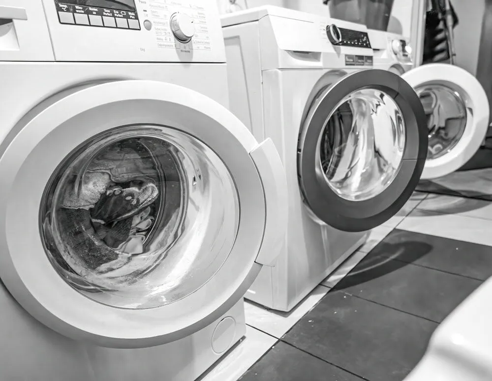 Two front-loading washing machines sit side by side in a North Attleboro laundry room. The black and white image reflects the surroundings, hinting at the need for reliable HVAC Contractor North Attleboro services to keep things running smoothly.