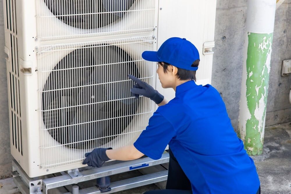 A technician in blue uniform and cap is kneeling and inspecting the fan of a large outdoor air conditioning unit, pointing at the metal grill—showcasing expert AC Services North Attleboro, MA.