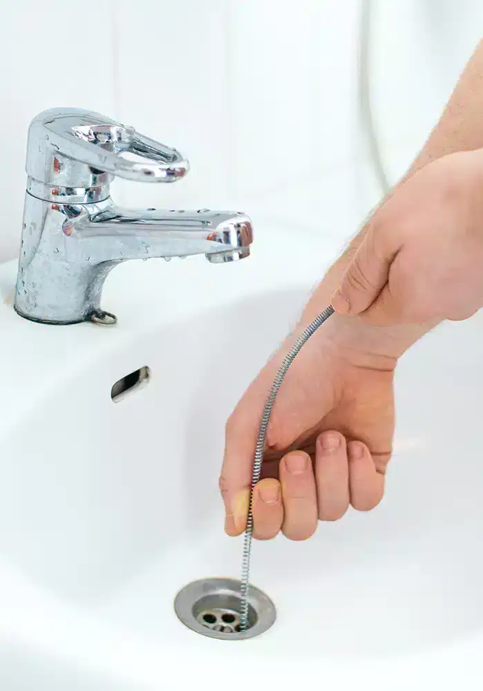 A person’s hand uses a flexible drain snake to unclog a bathroom sink, with a silver faucet and drain visible—professional tips often recommended by HVAC Contractor North Attleboro experts for routine home maintenance.