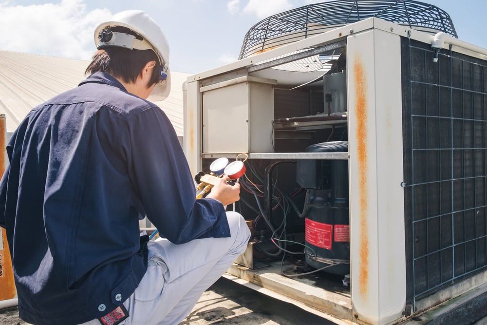 A technician wearing a hard hat and safety glasses checks gauges while inspecting an outdoor air conditioning unit on a rooftop, ensuring top-quality AC Services North Attleboro for reliable cooling performance.