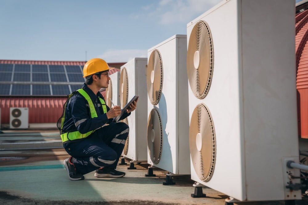 A worker wearing a hard hat, safety vest, and uniform inspects and takes notes on a tablet near several large outdoor air conditioning units on a rooftop for an HVAC Contractor North Attleboro, MA.