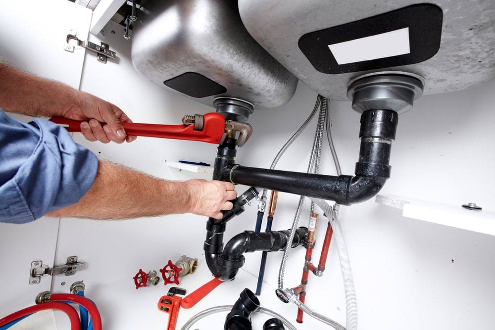 A person uses a red pipe wrench to tighten a black pipe under a double kitchen sink, surrounded by plumbing fittings and tools—typical of work done by an HVAC Contractor North Attleboro, MA.