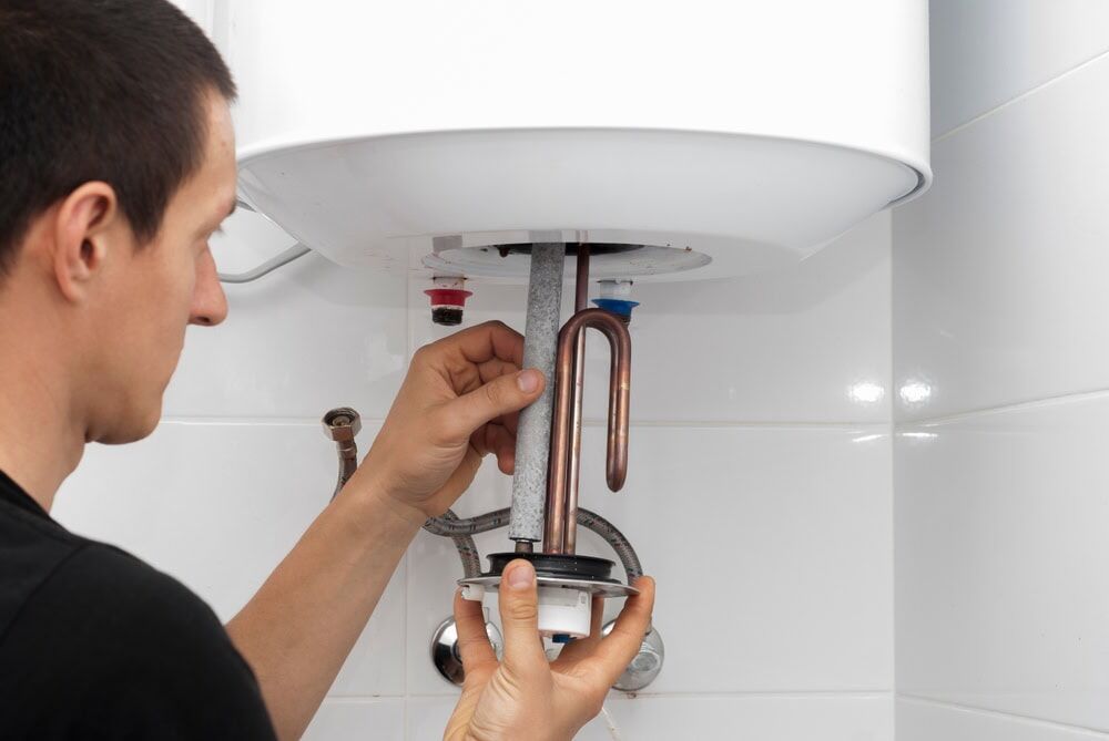 A person is repairing or installing a water heater in MA, holding a heating element and parts under the tank, with visible plumbing and pipes on a white tiled wall.