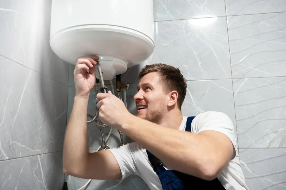 A smiling man in overalls uses a screwdriver to fix or install a white water heater mounted on a tiled bathroom wall, showcasing the expertise you can expect from AC Installation North Attleboro professionals.