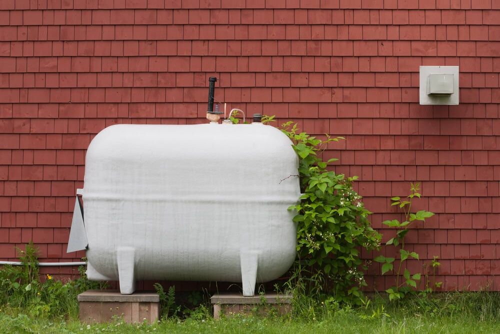 A large white propane tank stands on concrete blocks in front of a red shingled wall, surrounded by green grass—a typical sight for properties serviced by an HVAC Contractor North Attleboro, MA.