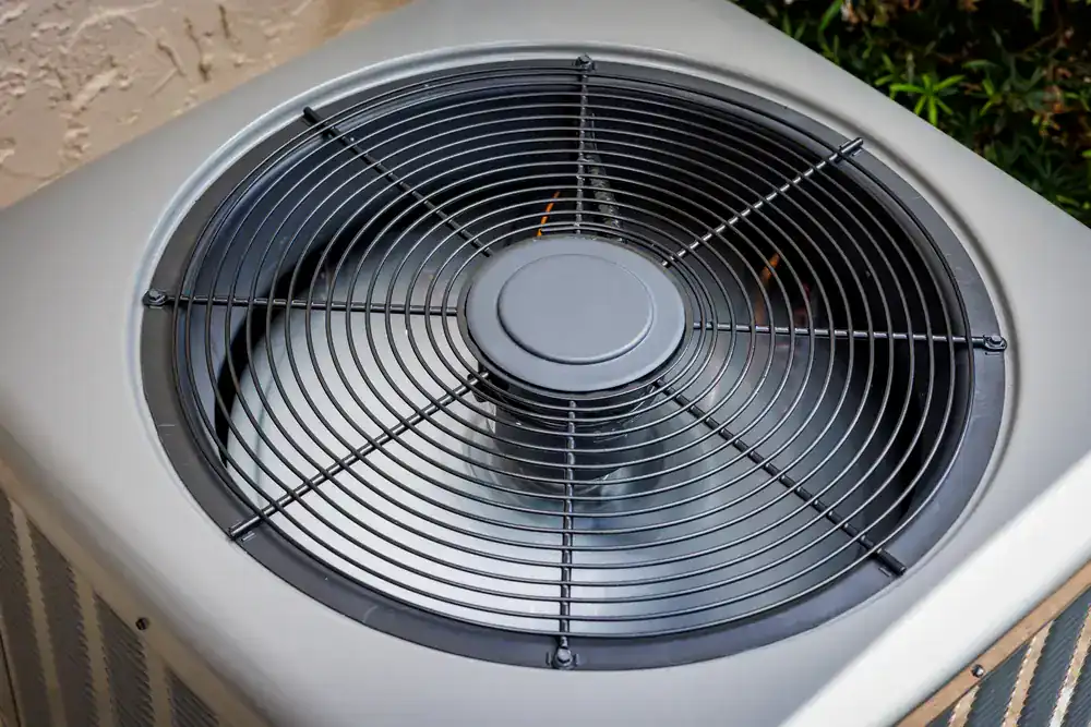 Close-up of the top view of an outdoor air conditioning unit by an HVAC Contractor North Attleboro, showing a large circular fan behind a protective metal grille. Some greenery and part of a building are visible in the background.