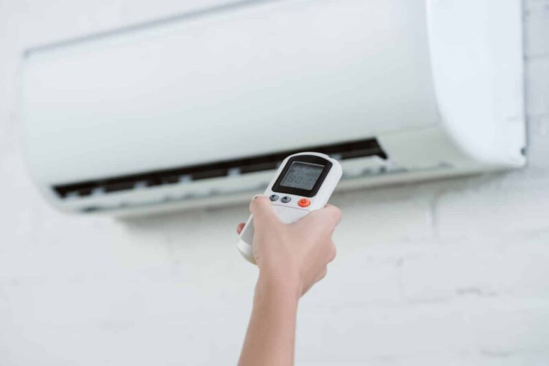 A person holds a remote control aimed at a split air conditioning unit mounted on a white brick wall. The remote's screen displays temperature settings.