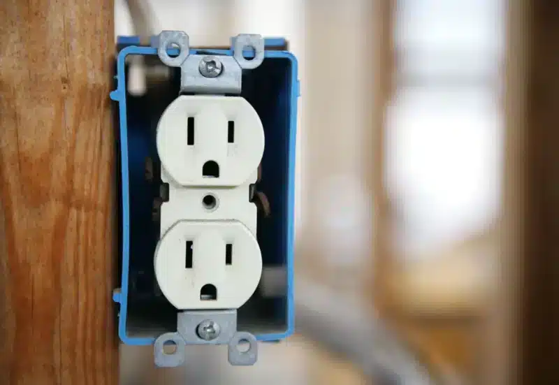 A close-up of a standard white electrical outlet mounted in a blue outlet box, attached to a wooden structure. The background is softly blurred, highlighting the outlet's details and wiring.