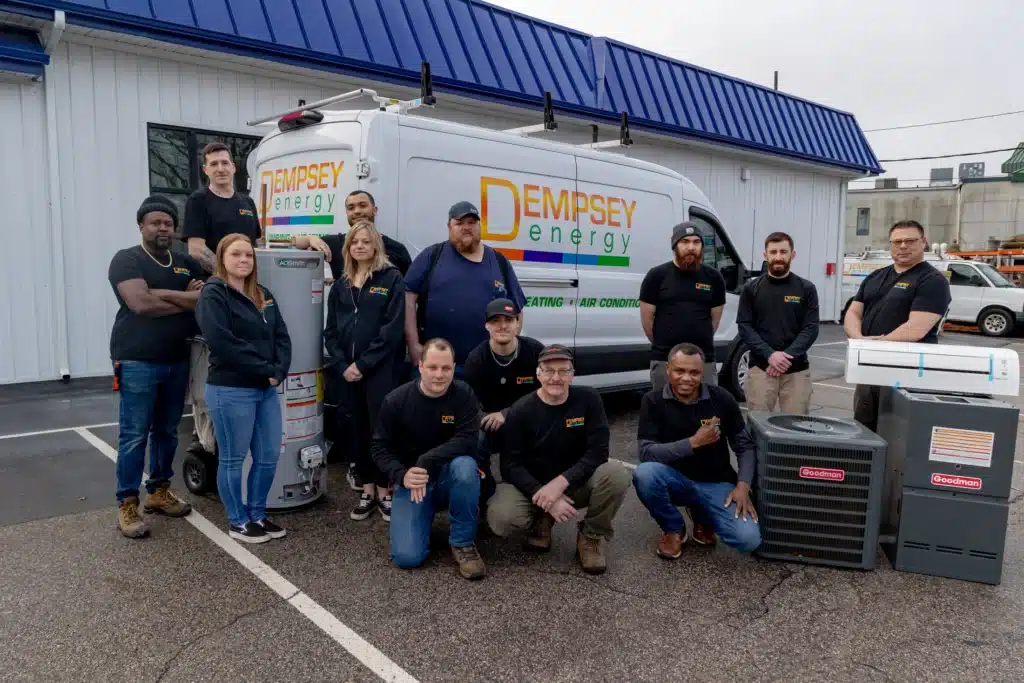 A group of twelve people wearing matching black shirts poses in front of a van with "Dempsey Energy" on it. The van is parked outside a building with a blue roof. Several air conditioning units are displayed in front.