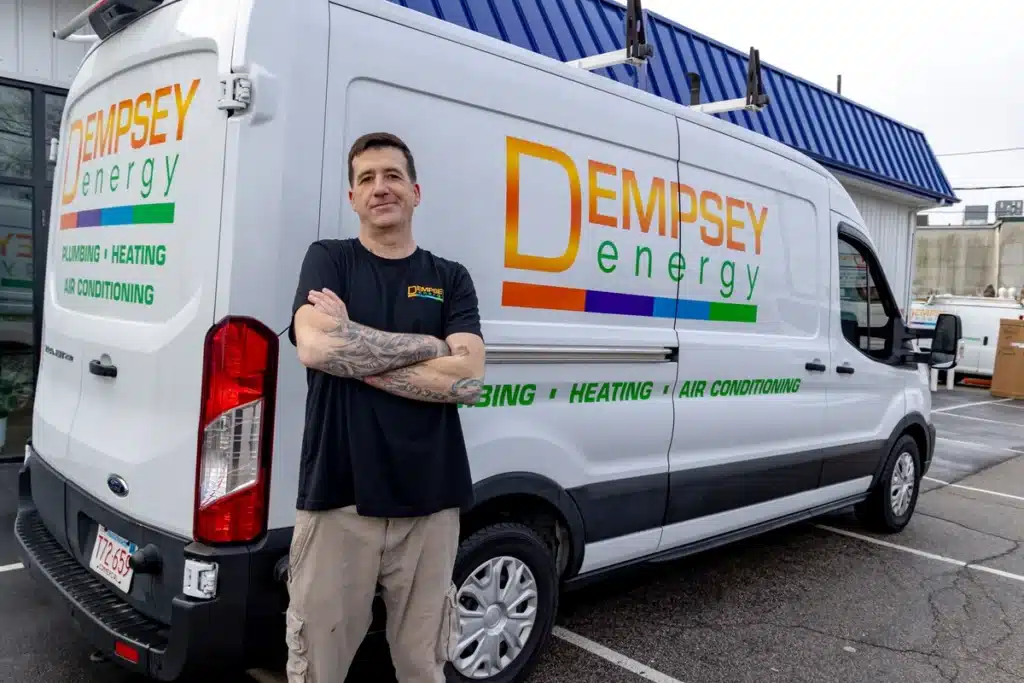 Man in a black shirt stands confidently with crossed arms in front of a white van bearing the "Dempsey Energy" logo. The van advertises plumbing, heating, and air conditioning services. A building is in the background.