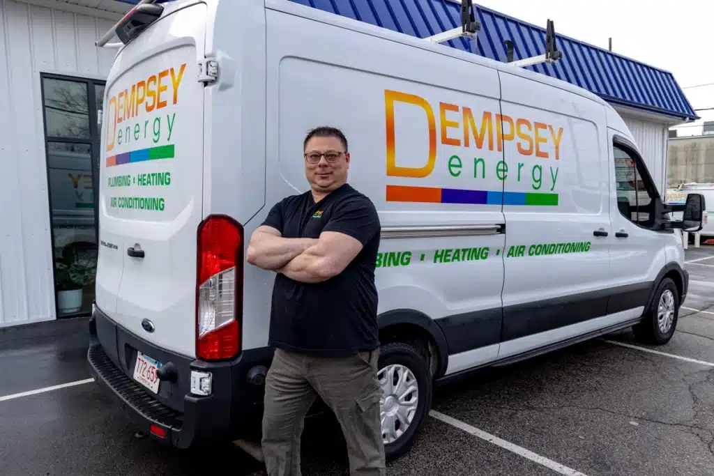 A man in a black shirt stands confidently with crossed arms in front of a white van labeled "Dempsey Energy". The van also displays "Plumbing, Heating, Air Conditioning" and is parked beside a building with a blue roof.