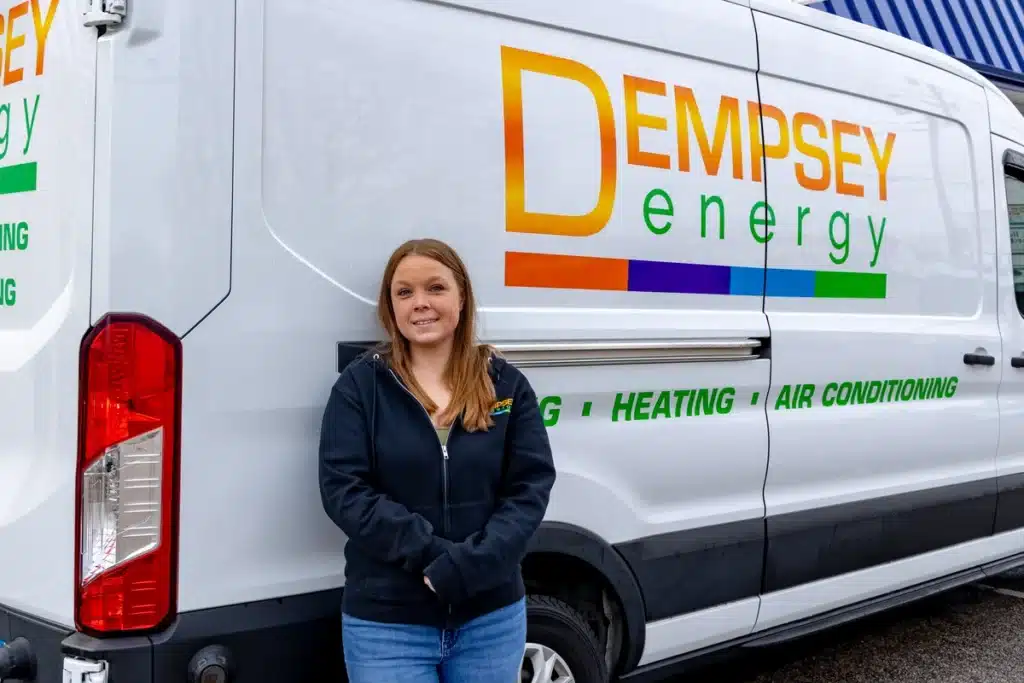 A woman stands in front of a white van displaying "DEMPSEY energy" with colorful lettering. The van lists services: plumbing, heating, and air conditioning. She wears a black zip-up jacket and jeans.