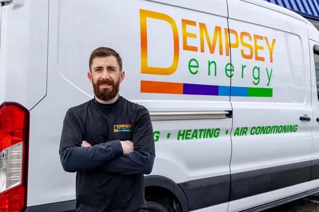 Man with a beard, arms crossed, stands in front of a white van with "Dempsey Energy" and colorful stripes on the side. The van also displays "Heating • Air Conditioning" services. The person wears a black shirt with the same company branding.