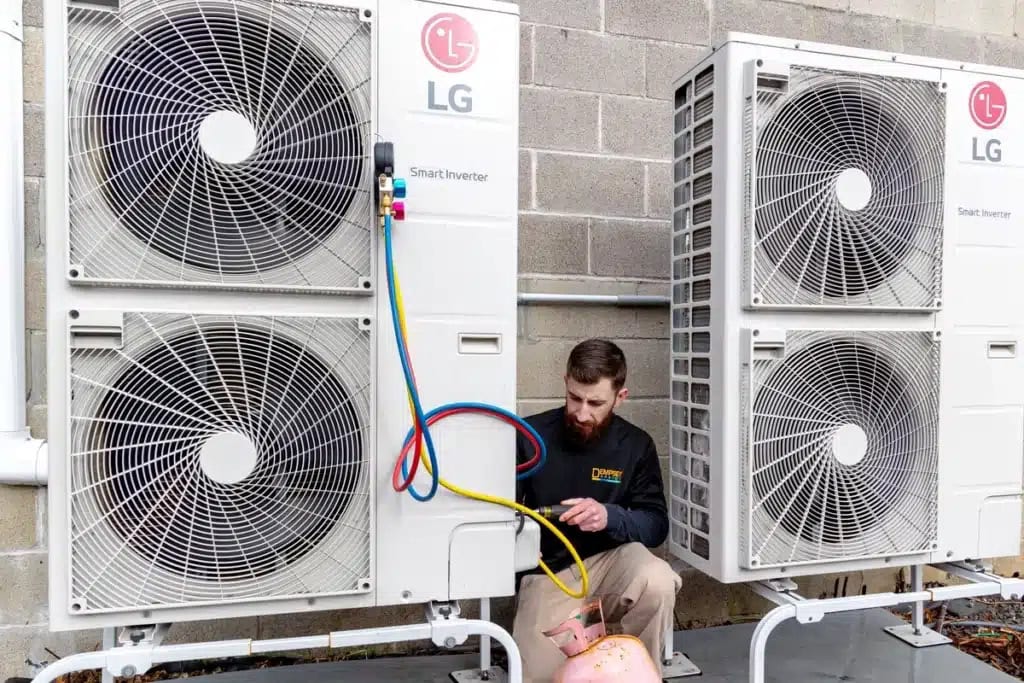 A technician with a beard is kneeling and working on a large LG air conditioning unit outdoors. He is using colorful hoses connected to the unit, with a pink tank resting nearby. The units are mounted against a brick wall.