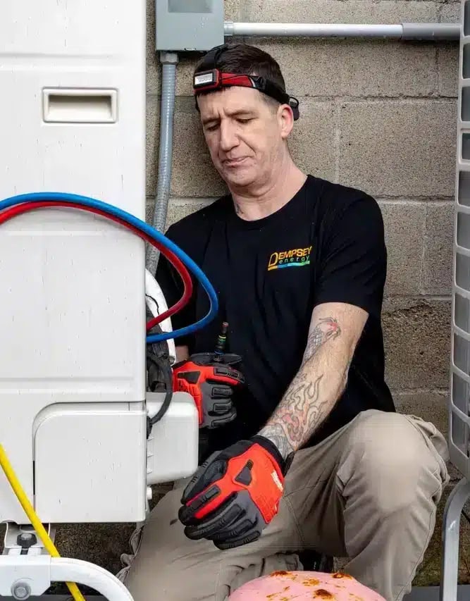A technician in a black shirt, beige pants, and red gloves works on an HVAC unit outdoors. He wears a headlamp and has tattoos on his left arm. Various tools and hoses are visible.