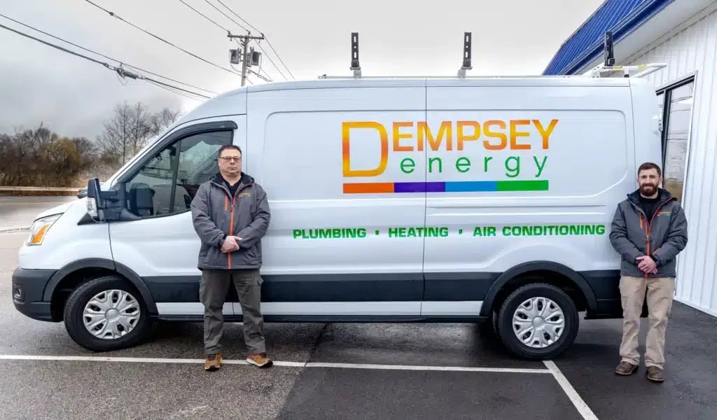 Two people stand beside a white service van with "Dempsey Energy" and "Plumbing - Heating - Air Conditioning" written on it. The van is parked outside a building on a cloudy day.
