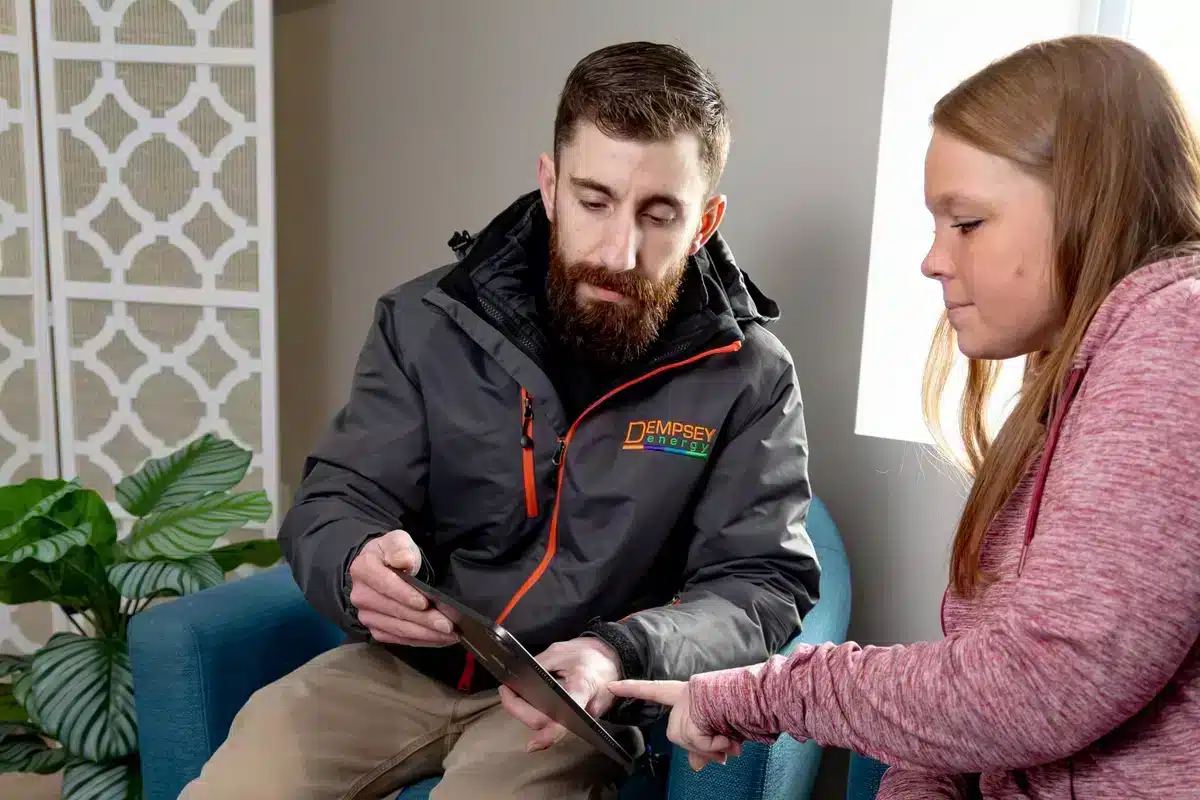 A man and a woman sit together in an office-like setting. The man, wearing a jacket with a visible logo, shows the woman something on a tablet. The woman points at the screen. They are seated near a decorative screen and a potted plant.
