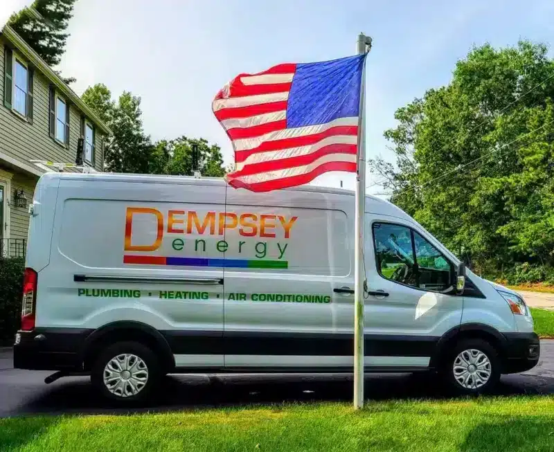 A white van with "Dempsey Energy: Plumbing, Heating, Air Conditioning" is parked on a driveway. An American flag waves in the foreground. Trees and a partial view of a house are in the background.