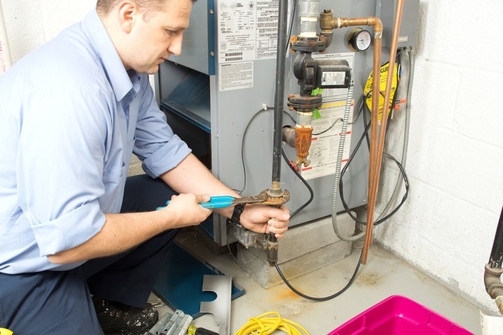 A man kneels on the floor using a wrench to tighten a pipe connected to a furnace in a utility room—typical work for an HVAC Contractor North Attleboro. Tools, wires, and a pink container are scattered nearby.